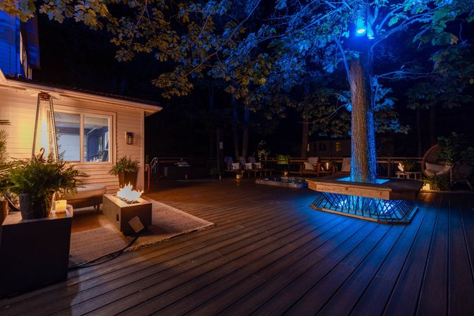 Nighttime backyard deck with wooden decking, blue uplighting on a large tree, glowing rectangular fire pit, potted plants and cozy outdoor seating