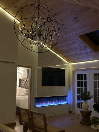 Cozy modern living room with wood-paneled vaulted ceiling, crystal orb chandelier, LED accent lighting, wall-mounted TV above a long blue-lit electric fireplace with marble surround, French doors, and a peek into a tufted-bed bedroom.