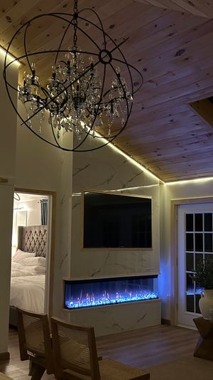 Cozy modern living room with wood-paneled vaulted ceiling, crystal orb chandelier, LED accent lighting, wall-mounted TV above a long blue-lit electric fireplace with marble surround, French doors, and a peek into a tufted-bed bedroom.