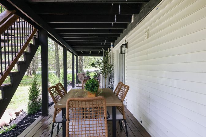 Covered wooden porch deck with outdoor dining: a rectangular wooden table, wicker chairs, potted greenery and seating beneath an exterior staircase overlooking a leafy backyard