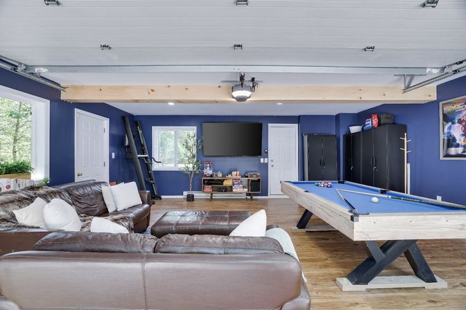 Finished garage rec room with blue walls, leather sectional, wall-mounted TV, blue-felt pool table, wood floors and black storage cabinets