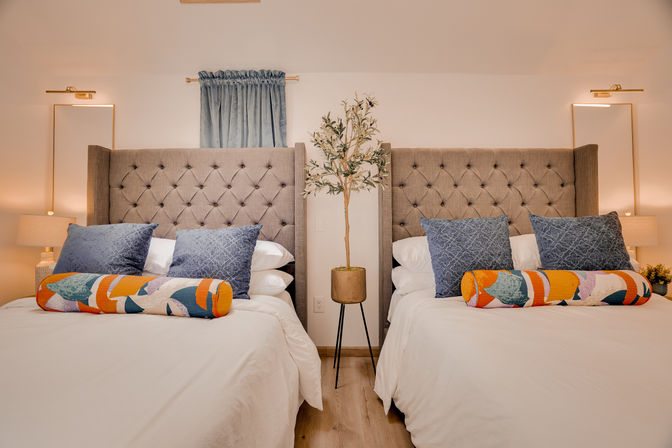 Cozy modern twin bedroom interior with tufted gray upholstered headboards, blue accent pillows and colorful bolster cushions, potted olive tree on a tripod stand between beds, warm bedside lamps and neutral decor.