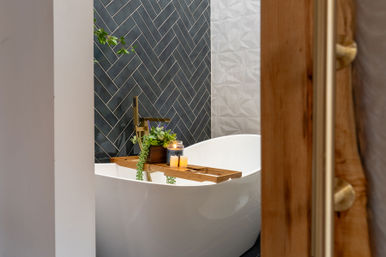 Spa-like modern bathroom with white freestanding tub, dark herringbone tile accent wall, brass floor faucet and a wooden bath tray holding candles and potted greenery.