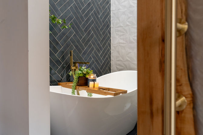 Spa-like modern bathroom with white freestanding tub, dark herringbone tile accent wall, brass floor faucet and a wooden bath tray holding candles and potted greenery.