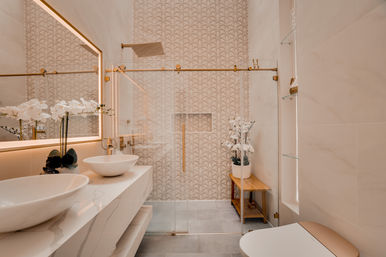 Spa-like modern bathroom with marble vanity and vessel sinks, backlit mirror, glass walk-in shower with gold hardware and geometric patterned tile accent wall, wooden bench and white orchids in soft beige tones.