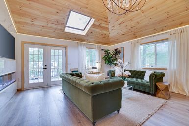 Cozy rustic-modern mountain cabin living room with vaulted pine ceiling and skylight, twin green velvet Chesterfield sofas, glass French doors to a wooded deck, hardwood floors, chandelier, indoor plant and white orchid centerpiece.