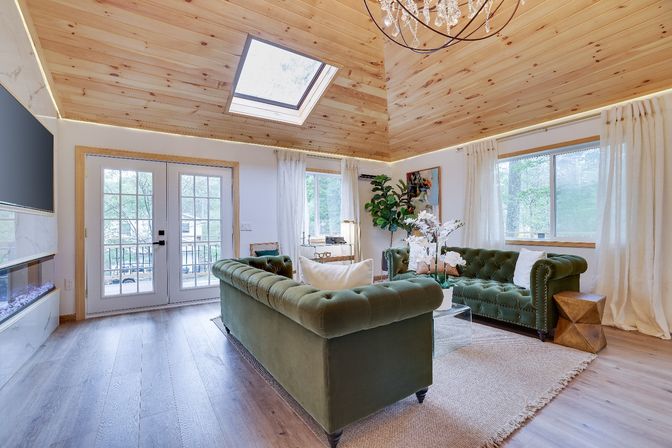 Cozy rustic-modern mountain cabin living room with vaulted pine ceiling and skylight, twin green velvet Chesterfield sofas, glass French doors to a wooded deck, hardwood floors, chandelier, indoor plant and white orchid centerpiece.