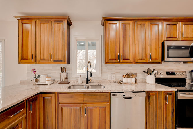 Bright modern kitchen interior with warm wooden cabinets, white marble-look quartz countertops, undermount sink and black pull-down faucet, stainless dishwasher and microwave, knife block, utensil holders and small potted orchid on the counter.