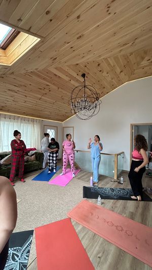 Cozy loft yoga session: group of women in pajamas and loungewear on colorful mats in a bright room with a wood-paneled vaulted ceiling, skylight and chandelier.