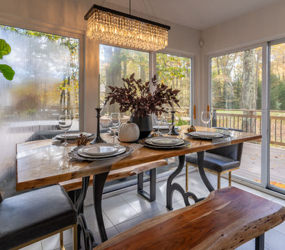 Modern rustic dining room with live-edge wood table set for four, crystal chandelier, leather chairs and bench, large windows and sliding glass doors overlooking an autumn wooded backyard and deck with barbecue