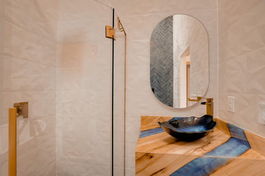 Sleek contemporary bathroom with glass shower and gold hardware, oval mirror, black vessel sink on a wood-and-blue resin countertop, and textured white tile walls