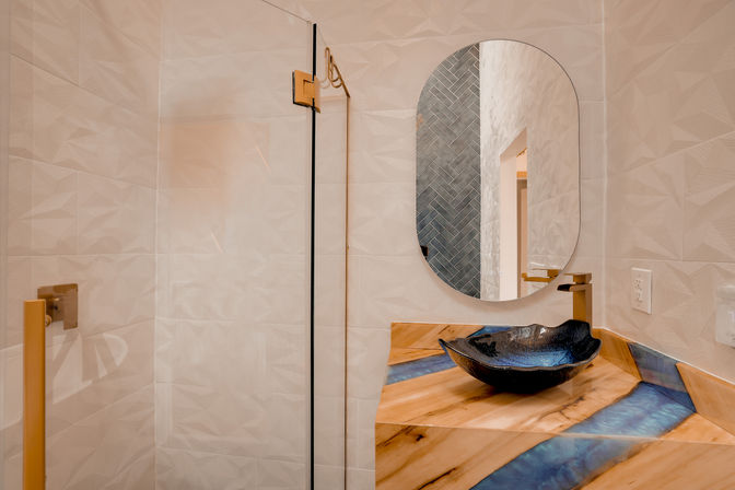 Sleek contemporary bathroom with glass shower and gold hardware, oval mirror, black vessel sink on a wood-and-blue resin countertop, and textured white tile walls