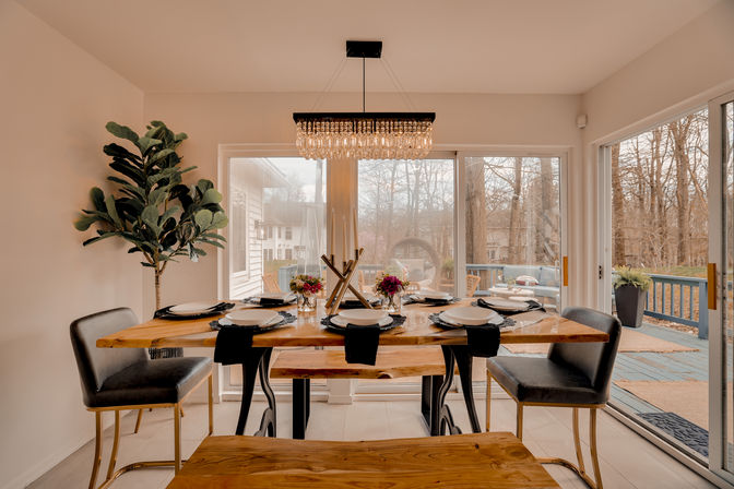 Modern dining room with live-edge wood table set for six, black velvet chairs, crystal chandelier and centerpiece, and large sliding glass doors opening to a suburban backyard deck and wooded view.