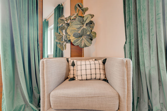 Cozy beige armchair with plaid tassel pillow framed by green velvet curtains and a fiddle-leaf fig plant in a modern living room nook