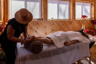 Relaxing foot massage in a cozy wood‑paneled spa room — therapist working on a client on a white‑towel treatment table with candles, red flowers, and soft natural light.