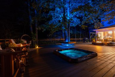 Nighttime backyard deck in a wooded setting with a glowing hot tub, fire pit, cozy outdoor seating and dramatic blue uplighting on trees