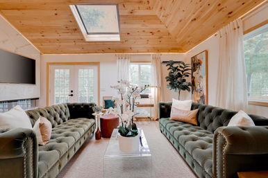 Sunny cozy living room with vaulted pine ceiling and skylight, twin tufted green velvet sofas facing a clear acrylic coffee table topped with white orchids, French doors and large windows bringing in natural light