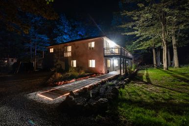 Cozy two-story house at night with warm interior lights, illuminated stone walkway edged in string lights, second-floor deck and tall trees in a wooded yard