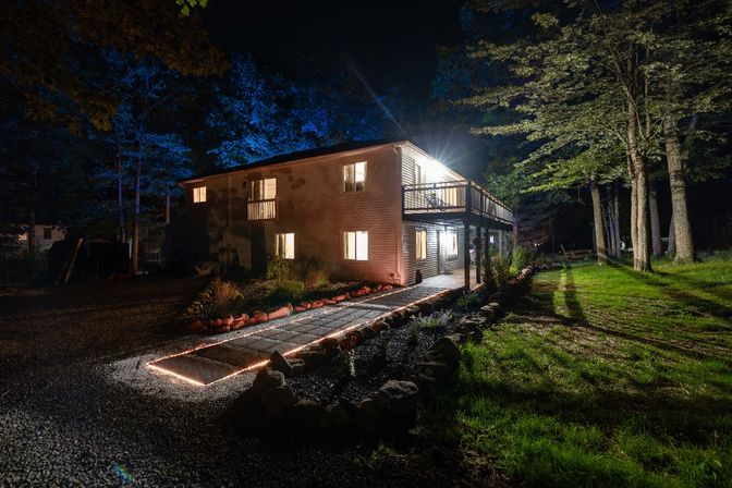 Cozy two-story house at night with warm interior lights, illuminated stone walkway edged in string lights, second-floor deck and tall trees in a wooded yard