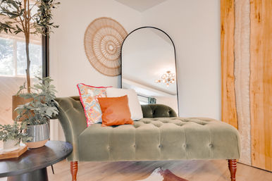 Cozy boho-chic living room corner with an olive-green tufted chaise lounge, colorful patterned and leather pillows, arched black-framed floor mirror, round woven wall decor, potted plants, wooden side table and warm natural light.