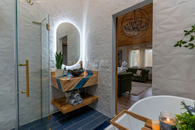 Bright modern bathroom with geometric white tile, glass shower with brass hardware, floating wood vanity with blue resin inlay and oval LED mirror, freestanding tub with candle and tray, and an open doorway showing a cozy living room with a green velvet sofa.