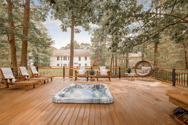 Spacious wooden backyard deck with built-in hot tub, multiple lounge chairs and a hanging wicker egg chair under tall trees, overlooking a suburban house and wooded yard.