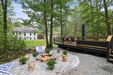 Leafy backyard with a stone fire pit burning in a paved patio, log stools and potted ferns, adjacent raised wooden deck with outdoor seating and a white two-story house in the background