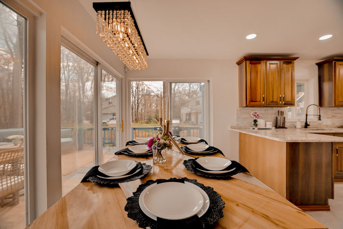 Open-plan modern kitchen and dining area with a wooden island table set for six on black placemats and white plates, a floral centerpiece, rectangular crystal chandelier overhead, and sliding glass doors to a backyard deck and trees.
