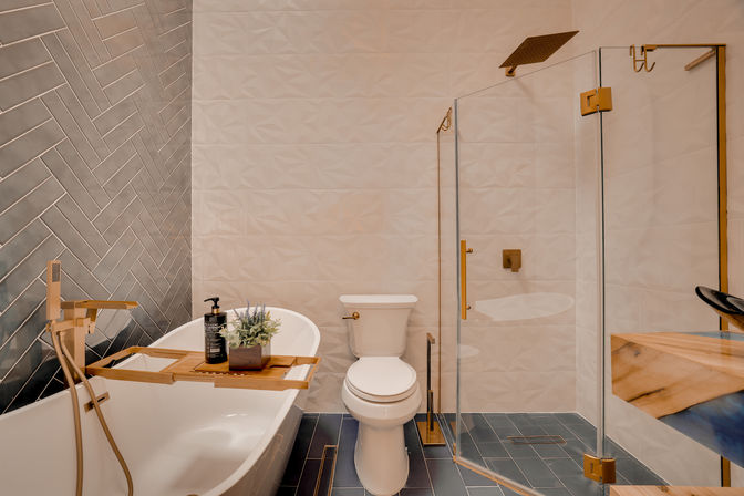 Modern luxury bathroom with freestanding white tub and wooden caddy, gold floor-mounted faucet, white toilet, glass-enclosed shower with gold fixtures, textured white walls and dark herringbone tile accent.