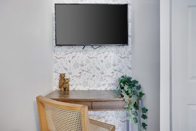 Cozy compact workspace with wall-mounted TV, wooden floating desk, cane chair and trailing ivy against whimsical animal-print wallpaper.