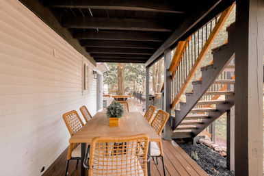 Cozy covered backyard deck dining area with a long wooden table, wicker chairs and a small potted centerpiece, beside open wooden stairs leading to an upper deck and tree-lined yard.