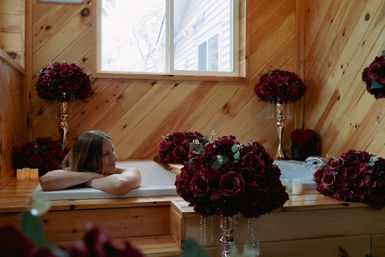 Woman relaxing in a wood-paneled bathtub surrounded by deep red rose bouquets, candles, and a window — cozy romantic spa-like scene.