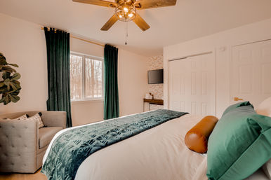 Cozy modern bedroom with white bedding, green velvet throw and curtains, leather bolster pillow, tufted armchair, ceiling fan, wall-mounted TV and window view of snowy woodland