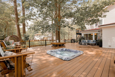 Spacious residential wooden deck with inset hot tub, large shade trees and circular bench, cozy outdoor sofa and swing chairs beside a white house — a relaxing backyard patio.