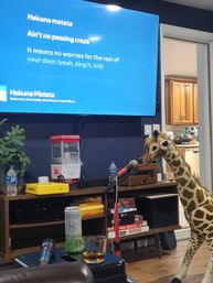Living room karaoke scene with TV displaying "Hakuna Matata" lyrics, microphone on a stand, a large plush giraffe leaning in to 'sing', popcorn maker, drinks and stacked board games on a media console.