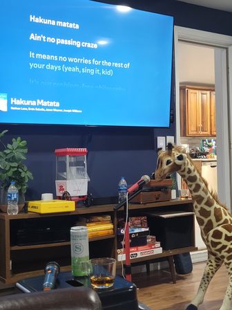 Living room karaoke scene with TV displaying "Hakuna Matata" lyrics, microphone on a stand, a large plush giraffe leaning in to 'sing', popcorn maker, drinks and stacked board games on a media console.