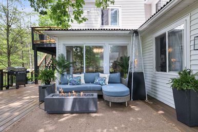 Cozy backyard wooden deck patio with a blue cushioned sectional and patterned pillows, modern rectangular fire pit, tall patio heater, potted plants, grill, and sliding glass doors on a white-sided house surrounded by trees.