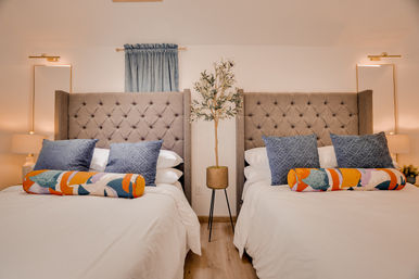 Cozy modern twin bedroom interior with tufted gray upholstered headboards, blue accent pillows and colorful bolster cushions, potted olive tree on a tripod stand between beds, warm bedside lamps and neutral decor.