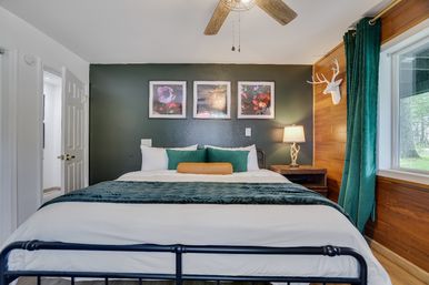 Cozy modern-rustic bedroom with dark green accent wall and king bed in white linens, teal throw and pillows, three framed floral prints above the headboard, wooden wall with faux deer head, bedside lamp, ceiling fan, and large window overlooking a wooded yard.