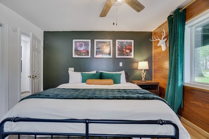 Cozy modern-rustic bedroom with dark green accent wall and king bed in white linens, teal throw and pillows, three framed floral prints above the headboard, wooden wall with faux deer head, bedside lamp, ceiling fan, and large window overlooking a wooded yard.