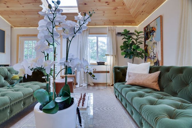 Cozy modern living room with wood-paneled vaulted ceiling and skylight, green tufted velvet sofas, white orchid centerpiece on a glass coffee table with a chess set, large window with light curtains and a leafy indoor plant.