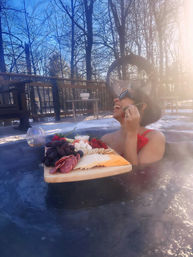 Person in red swimsuit and sunglasses relaxing in an outdoor hot tub on a snowy backyard deck with sun flare, holding a floating charcuterie board of cheeses, cured meats, grapes and strawberries.