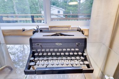Vintage Royal typewriter on a glass desk by a sunlit window overlooking a leafy suburban backyard