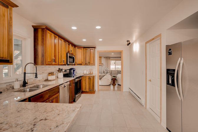 Spacious open-concept home kitchen with warm wood cabinets, granite countertops, stainless steel appliances, tile floor, recessed lighting and a view into a cozy living room