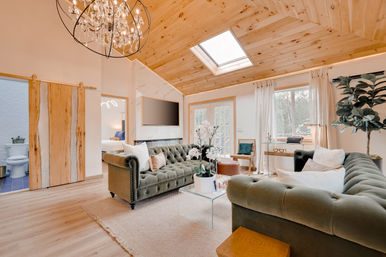 Bright, cozy living room with vaulted pine ceiling and skylight, crystal chandelier, matching olive-green tufted sofas around a glass coffee table with potted orchids, neutral rug, French doors, large windows, indoor plant and sliding wood barn door to bathroom.