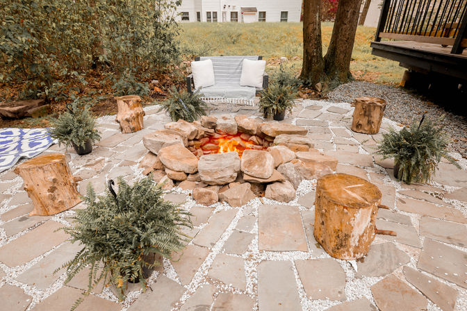 Cozy backyard fire pit patio with stacked-rock fire pit, flagstone pavers, log-stump seats, potted ferns, and a cushioned outdoor bench.
