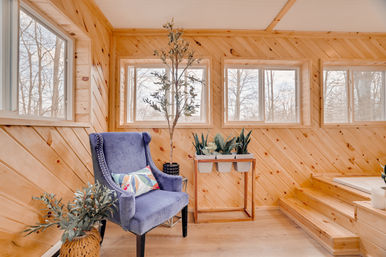 Cozy bright wood-paneled sunroom reading nook with a blue velvet wingback chair and colorful pillow, potted plants on a wooden stand, large windows and warm natural light.
