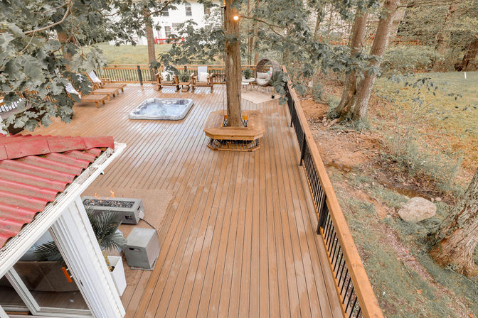 Aerial view of a spacious wooden backyard deck with a hot tub, lounge chairs, fire pit, wraparound tree bench and black metal railing, surrounded by trees and grassy yard.