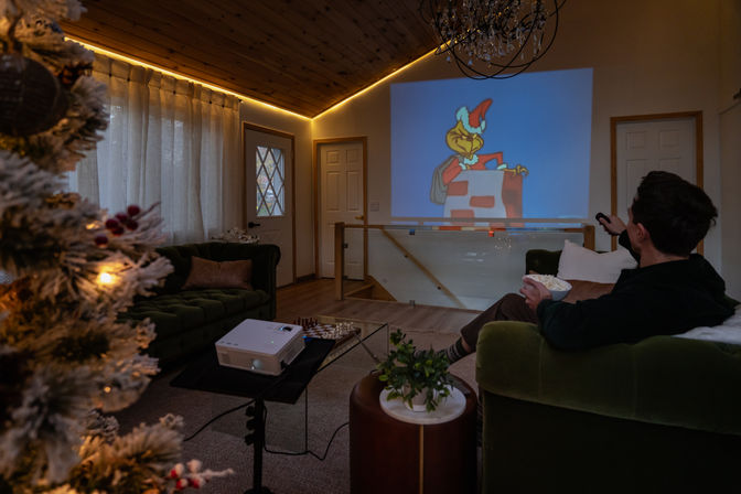 Cozy holiday living room and home theater—lit Christmas tree and warm LED trim, person on a green sofa with popcorn and remote watching a festive cartoon projected on the wall from a tabletop projector.