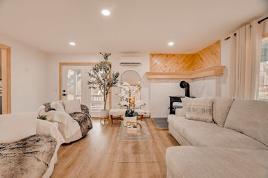 Cozy bright modern farmhouse living room interior with a neutral sectional and armchairs draped in faux-fur throws, glass coffee table with orchids, wood floors, potted tree, and a wood-paneled fireplace with a black stove.